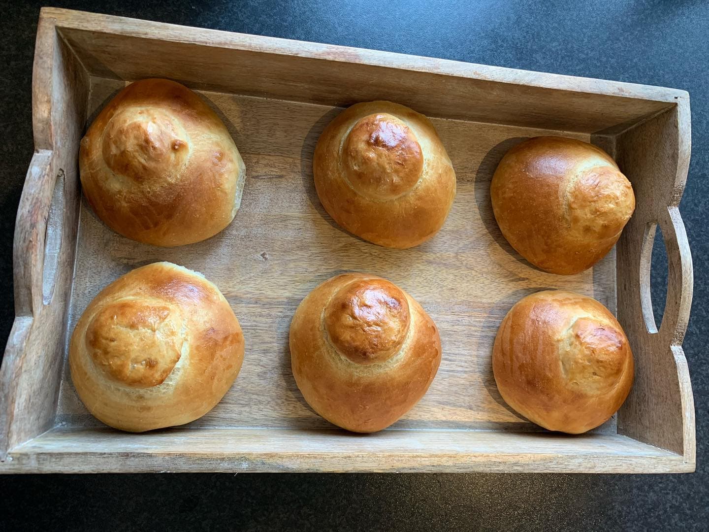 Six golden-brown brioche rolls with top knots arranged in a rustic wooden tray.