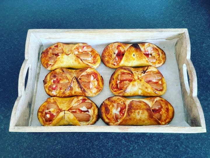 Six golden-brown sausage rolls with tomato sauce and cheese on a wooden serving tray.