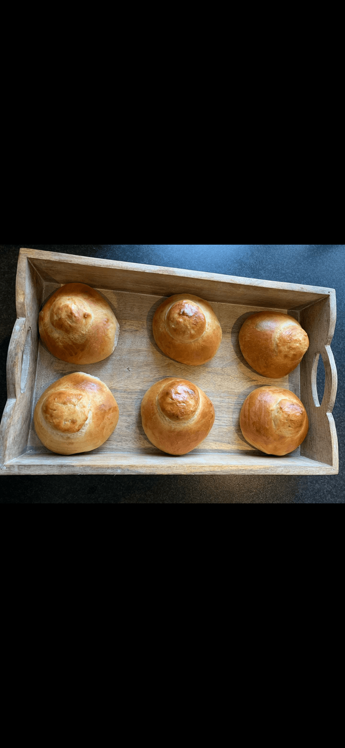 Six golden-brown brioche buns with top knots arranged inside a rectangular wooden serving tray.