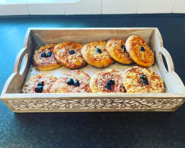 Eight mini pizzas with cheese and black olives arranged in a decorative carved wooden tray.